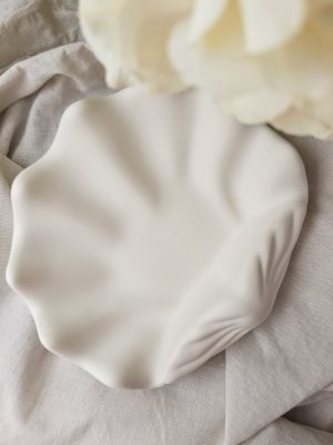 Weiße, muschelförmige Schmuckschale aus Raysin, von Hand gehalten vor beigem Stoffhintergrund mit Hortensien