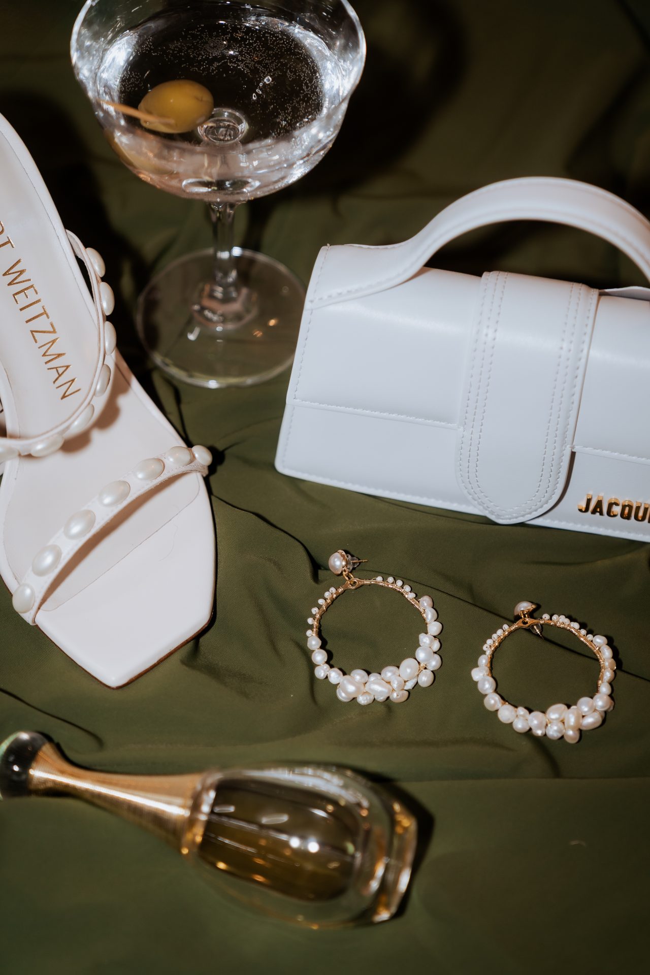 Auf einem grünen Stoff am Boden liegen ein weißer Brautschuh, eine kleine elegante Mini-Tasche, auffällige Pearl Luxe Statement-Ohrringe, ein Glas Martini mit Oliven und ein Flakon J’adore Parfum.
