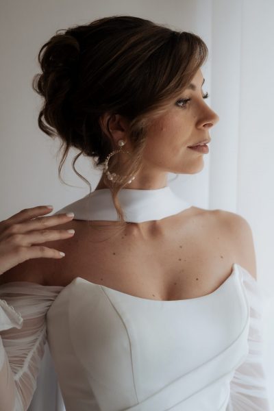 Braut im weißen Kleid steht vor einem großen Fenster, Licht strahlt von hinten, sie blickt nach links und trägt auffällige Pearl Luxe Statement-Ohrringe